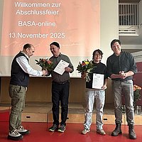 Im Audimax der ASH Berlin stehen vier Personen nebeneinander. Sie schauen in die Kamera und halten Blumen in der Hand. 