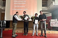 Im Audimax der ASH Berlin stehen vier Personen nebeneinander. Sie schauen in die Kamera und halten Blumen in der Hand. 