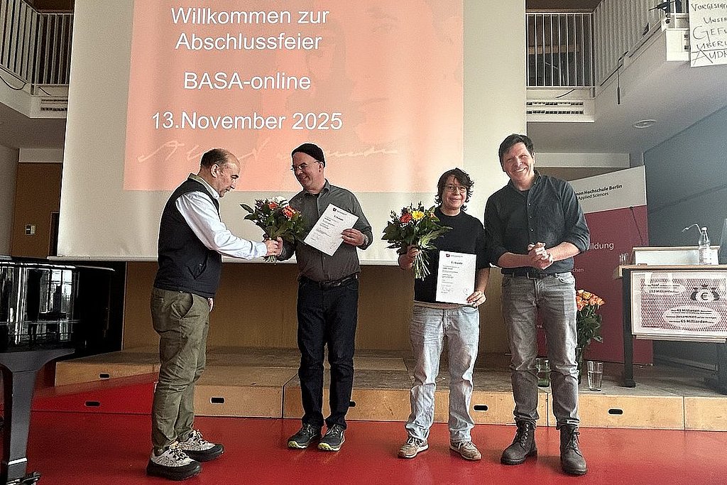 Im Audimax der ASH Berlin stehen vier Personen nebeneinander. Sie schauen in die Kamera und halten Blumen in der Hand. 