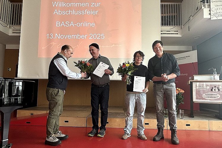 Im Audimax der ASH Berlin stehen vier Personen nebeneinander. Sie schauen in die Kamera und halten Blumen in der Hand. 