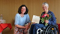 Bettina Völter sitzt neben Sigrid Arnade auf der Bühne im ASH-Audimax. Sigrid Arnade hält die Urkunde und einen Blumenstrauß in den Händen. 