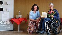 Bettina Völter sits next to Sigrid Arnade on the stage in the ASH Audimax. Sigrid Arnade holds the certificate and a bouquet of flowers in her hands.