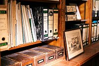 Blick in das Regal eines Archives. Alte Mappen und Ordner stehen und liegen auf Regalbrettern, darunter kleine Holzkästen. An dem Regal lehnt ein altes Foto in einem Bilderrahmen.