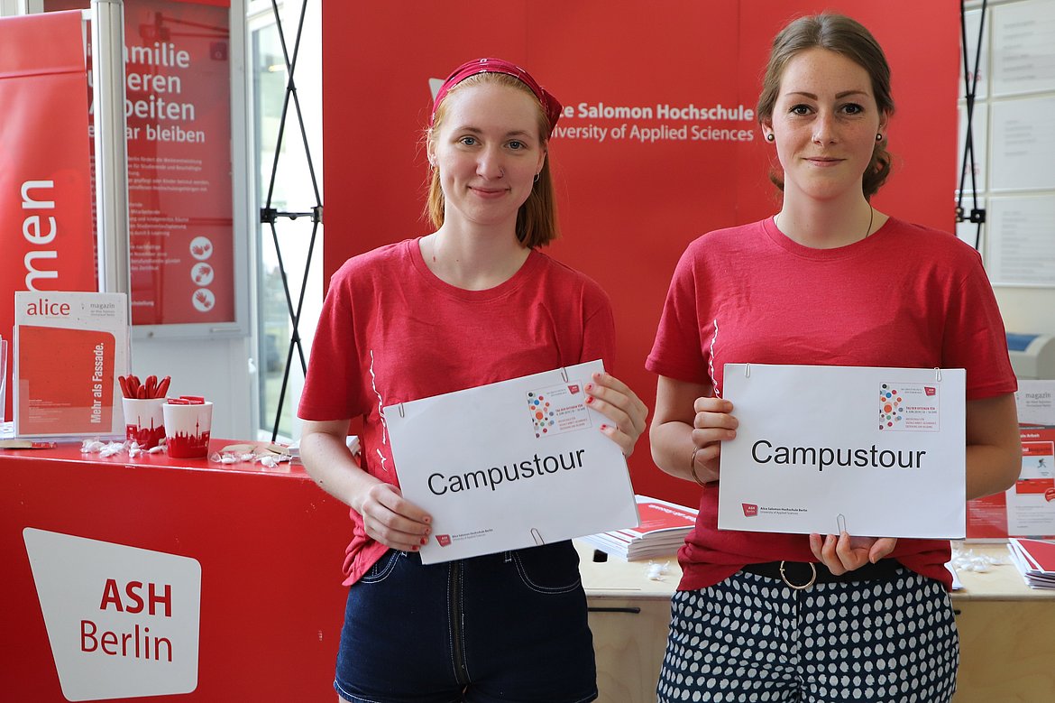 Zwei Studentinnen* zeigen an der Hochschule das Schild "Campustour"