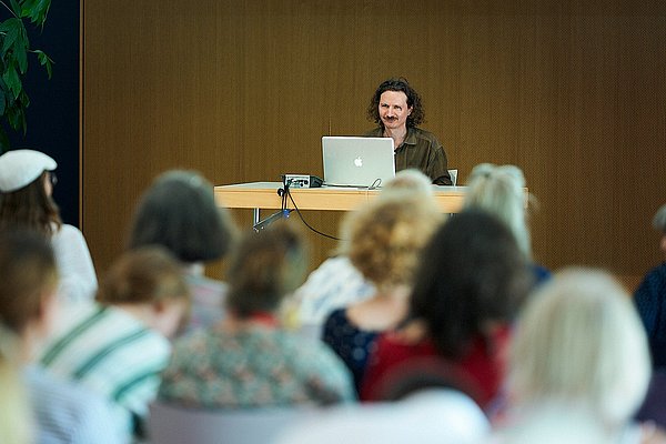 Der ukrainische Künstler Anatoly Belov auf der Bühne des ASH-Audimax. Er sitzt an einem Tisch, vor ihm steht ein Laptop.