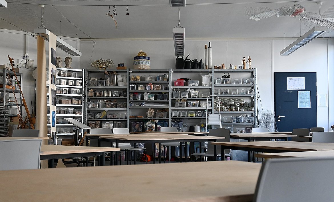 Es ist der Raum 118 zu sehen, mit Blick auf die Materialregale die hinter den Tischen mit Holzplatten stehen.