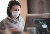 Eine Frau mit Mund-Nasenschutz im Gesicht sitzt an einem Laptop und arbeitet