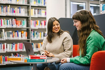 Zwei Studierende in der Bibliothek, vor ihnen auf dem Tisch Bücher und ein aufgeklappter Laptop, sie unterhalten sich.