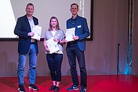 Auf dem Foto sind 3 Personen zu sehen die nebeneinander stehen und in die Kamera lächeln. Sie halten eine Urkunde in der Hand.