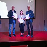 Auf dem Foto sind 3 Personen zu sehen die nebeneinander stehen und in die Kamera lächeln. Sie halten eine Urkunde in der Hand.