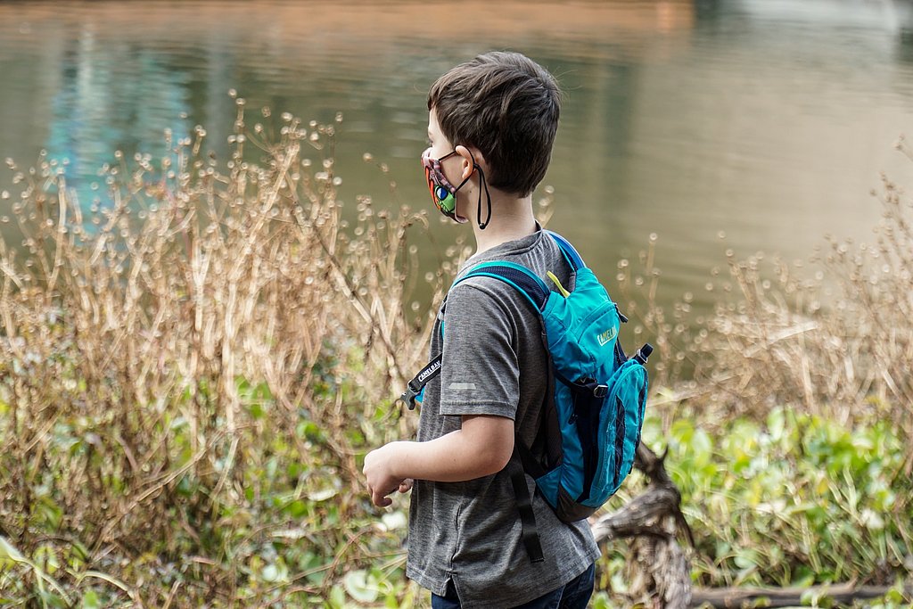 EIn Junge läuft an einem See vorbei, trägt einen Rucksack und eine Maske