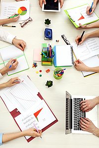 Vogelperspektive auf einen großen Schreibtisch, an dem arbeitende Menschen sitzen, wobei nur deren Hände abgebildet sind. Die Menschen schreiben am Laptop oder machen sich per Hand Notizen.