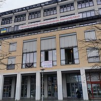 Auf dem Foto ist das Hauptgebäude der ASH Berlin zu sehen. Vor dem Fenster des Audimax ist ein Transparent zu sehen auf dem steht "Die Hochschule ist besetzt".