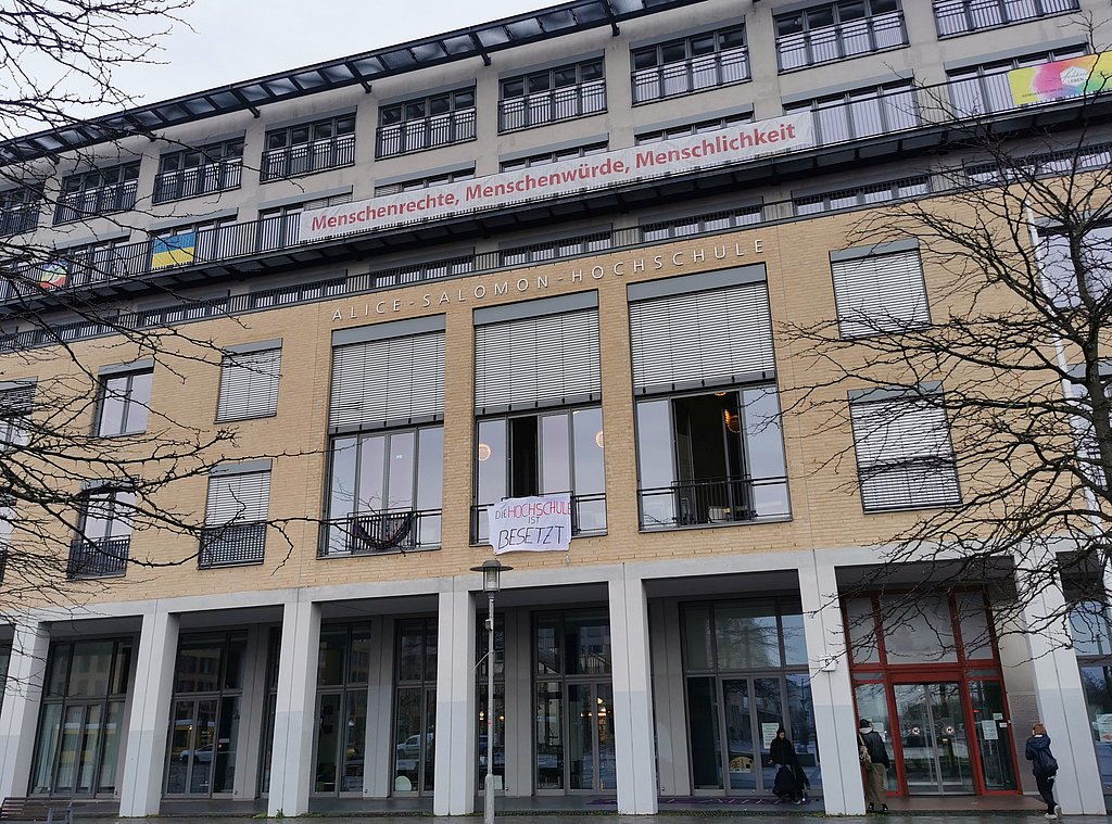 Auf dem Foto ist das Hauptgebäude der ASH Berlin zu sehen. Vor dem Fenster des Audimax ist ein Transparent zu sehen auf dem steht "Die Hochschule ist besetzt".