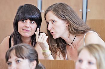 Zwei Studentinnen im Hörsaal, die eine flüstert der anderen etwas ins Ohr