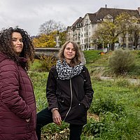 Ein Foto von Seba Abudaqa und Tom Kellner. Die beiden Frauen stehen am grünen Ufer eines Flusses, im Hintergrund sind eine Brücke und Wohnhäuser erkennbar