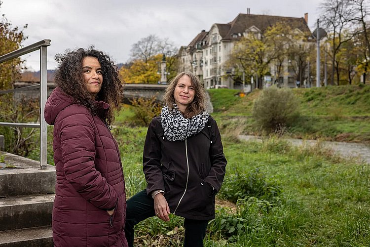 A photo by Seba Abudaqa and Tom Kellner. The two women are standing on the green banks of a river, with a bridge and residential buildings visible in the background