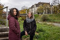 Ein Foto von Seba Abudaqa und Tom Kellner. Die beiden Frauen stehen am grünen Ufer eines Flusses, im Hintergrund sind eine Brücke und Wohnhäuser erkennbar