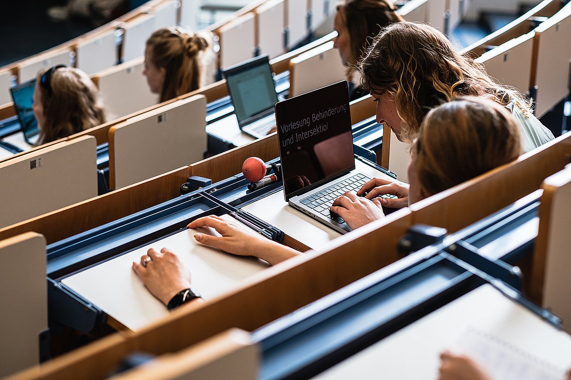 In einem Hörsaal beugt sich eine Person mit lockigen Haaren dicht über ein Laptop. Neben dem Laptop liegt ein Blindenlangstock. Im Hörsaal sitzen noch andere Studierende.