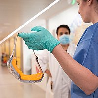 In einem Krankenhausflur: Pflegekraft hält Augenschutzmaske gegenüber anderem Mediziner, der Mundschutz trägt