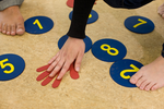 Auf dem Fußboden befinden sich runde Karten aus Karton mit Zahlen darauf sowie eine Karte in Form einer Hand. Eine Person beugt sich, um ihre Hand auf die Handkarte zu legen. 
