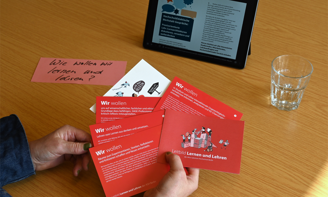 Postkarten-Set vom Leitbild Lernen und Lehren aufgefächert in der Hand. Im Hintergrund eine Karte mit Text: Wie wollen wir lernen und lehren? Dazu ein tablet und ein Glas Wasser auf einem Tisch.