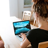 Das Bild zeigt eine junge Frau die an ihrem Laptop arbeitet. Vermutlich aus dem Home Office da sie an einem Esstisch sitzt.
