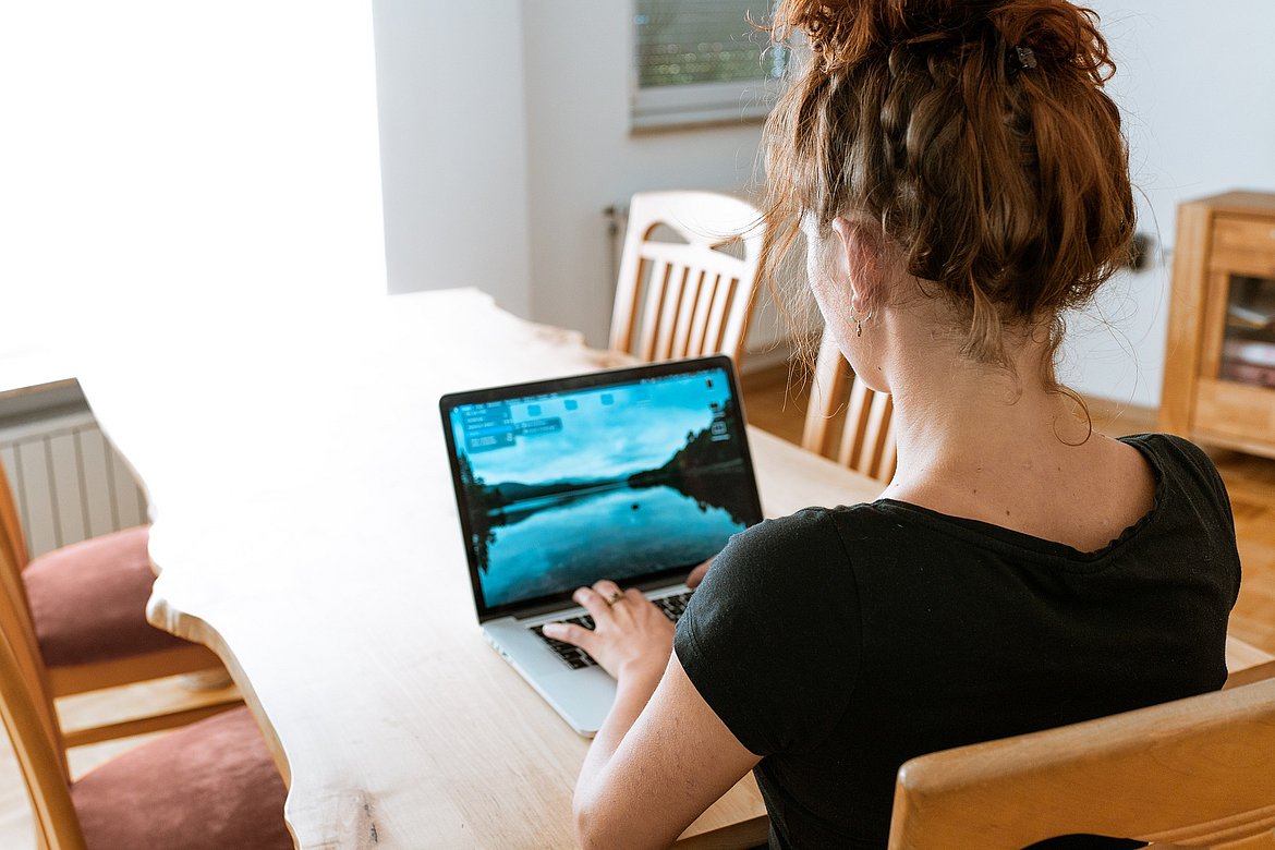 Das Bild zeigt eine junge Frau die an ihrem Laptop arbeitet. Vermutlich aus dem Home Office da sie an einem Esstisch sitzt.