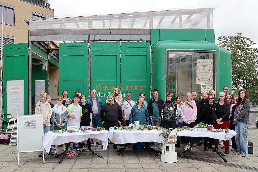 Vor dem Klassenzimmer der Zukunft, einem grünen Messebau, stehen Anwohner_innen und Schüler_innen hinter einem langen Tisch. Auf den weißen Tischdecken liegt Gemüse. 