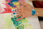 Eine Hand ist voll mit verschiedenen Fingermalfarben, mit denen ein Papier bunt bemalt wurde.