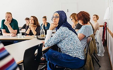 Studierende des Pre-Studies for Refugees Studiengangs sitzen am Tisch in einem Seminarraum der ASH Berlin Sie haben ihre Hände auf dem Tisch abgestützt und hören aufmerksam zu