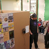 Aju John im Pavillion Klassenzimmer der Zukunft vor der ASH Berlin. Er hält ein Mikrofon in der Hand und spricht zu den Gästen die im Pavillion stehen. 