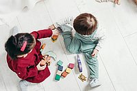 Das Foto zeigt zwei Kinder, die mit Bauklötzen und Meerestieren aus Holz spielen. Es ist aus der Perspektive eines nach unten blickenden Erwachsenen aufgenommen. Eines der Kinder trägt ein rotes Kleid, das andere eine türkisfarbene Latzhose.