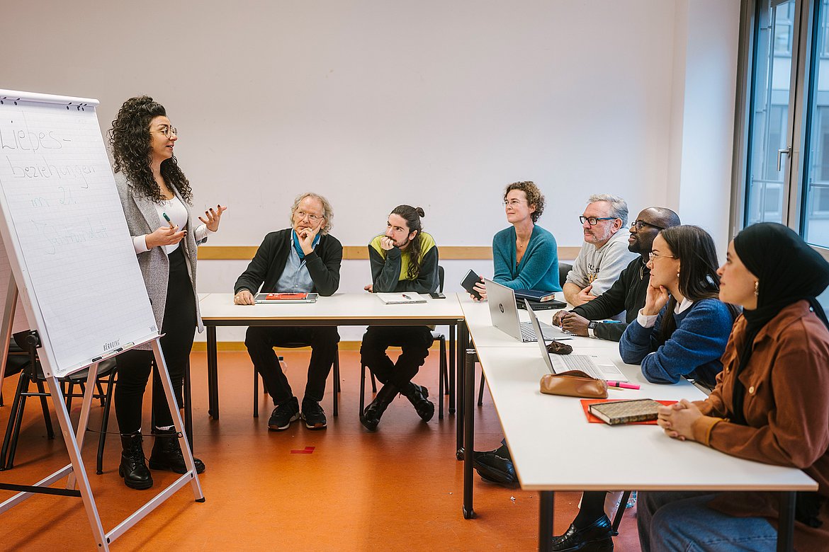 Studierende der ASH Berlin sitzen in einem Seminarraum der Hochschule und hören einem Vortrag zu. Eine Studierende steht neben einem Flipchart und hält einen Vortrag. 