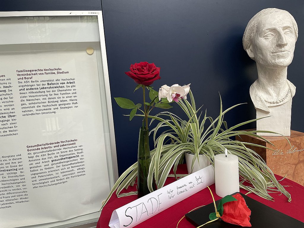 Auf dem Foto ist die Büste von Alice Salomon zu sehen, die sich im Foyer der ASH Berlin befindet, sowie ein Tisch mit Blumen, einer Trauerkerze und einem Kondolenzbuch