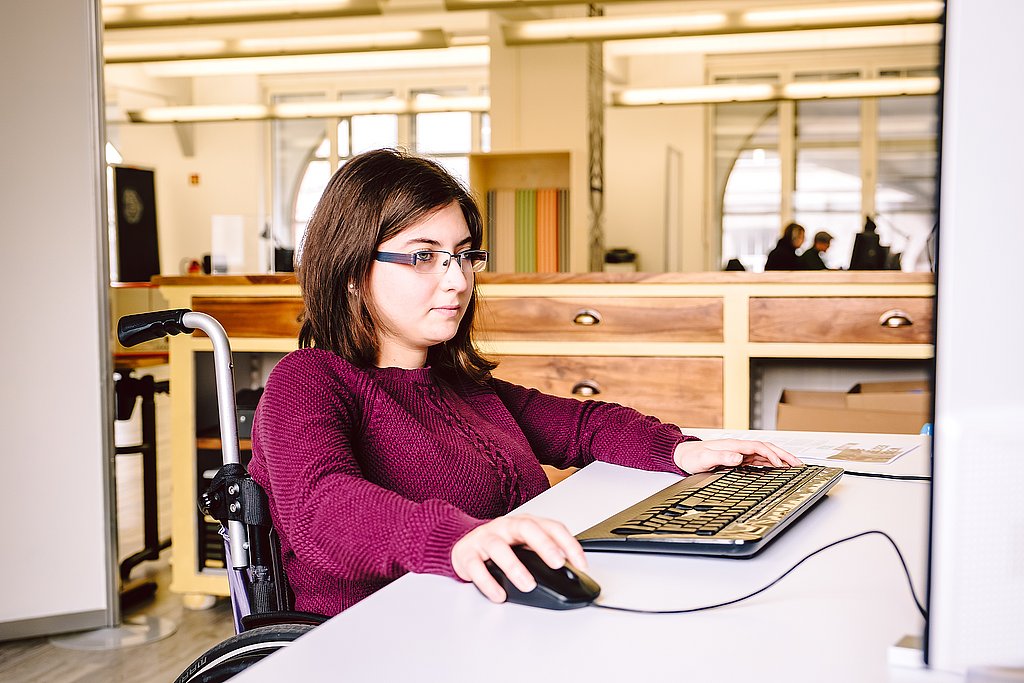 Eine Frau im Rollstuhl sitzt an einem Schreibtisch und arbeitet an ihrem Computer.
