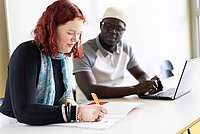 Zwei Studierende der ASH Berlin sitzen nebeneinander in einem Seminarraum. Eine Person schreibt etwas auf ein Papier, die andere Person schaut dabei zu.