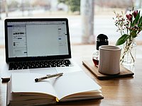 Laptop, in front of it an open notebook with a pen, on the edge a cup and a bouquet of flowers.