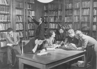 Historische schwarz-weiß-Aufnahme von Wissenschaftlerinnen und Wissenschaftlern, die sich in einer mit vielen Büchern gefüllten Bibliothek befinden und miteinander diskutieren.