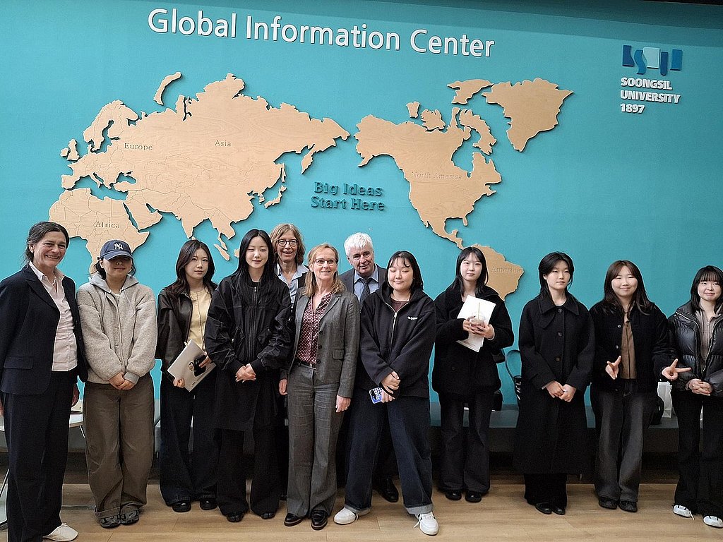 Die Delegation der ASH Berlin und Studierende der Soongsil University vor einem Wandbild mit einer Weltkarte. Über der Weltkarte steht der Schriftzug: Global Information Center