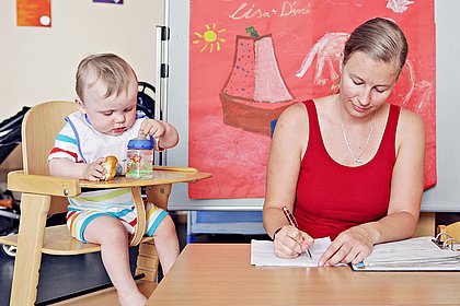 Eine Mutter arbeitet am Schreibtisch, daneben sitzt ihr Kind im Kinderstuhl und spielt mit seinem Fläschchen