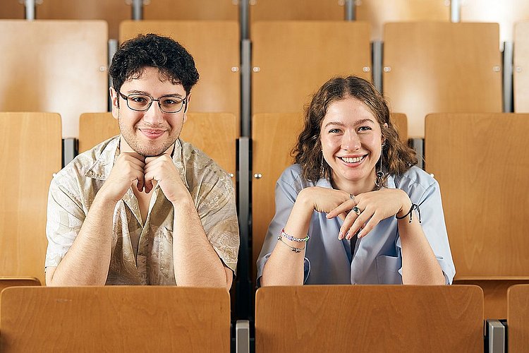 Zwei Studierende der ASH Berlin sitzen nebeneinander im Hörsaal und lächeln in die Kamera. 