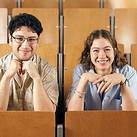 Zwei Studierende der ASH Berlin sitzen nebeneinander im Hörsaal und lächeln in die Kamera. 