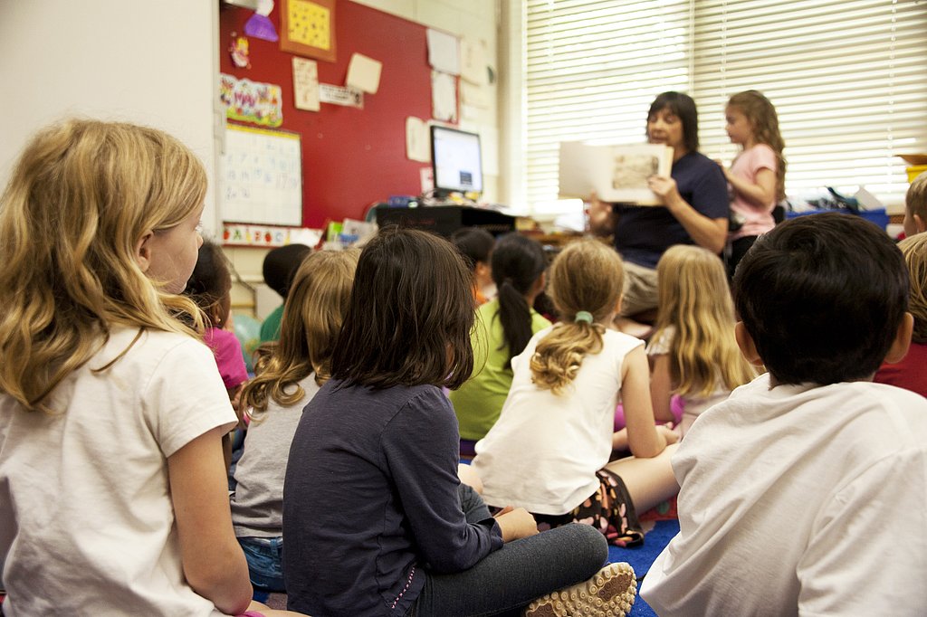 Auf dem Boden sitzende Kinder von hinten fotografiert - auf eine Lehrkraft mit Buch schauend