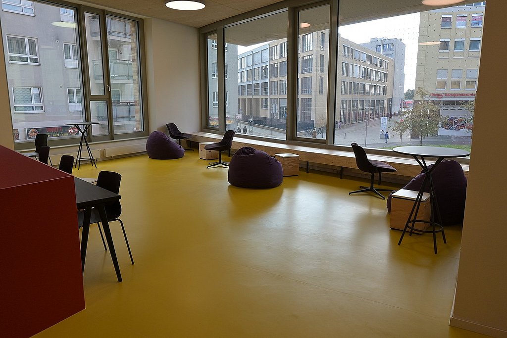 Eine Selbstlernfläche im Neubau, ausgestattet mit verschiedenen Sitzgelegenheiten. Durch große Fenster ist der Koschkaplatz und das Hauptgebäude der ASH Berlin zu sehen