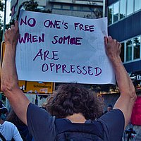 Demo von hinten fotografiert. Eine Person hält ein Schild hoch, auf dem steht: No one's free if some are oppressed.