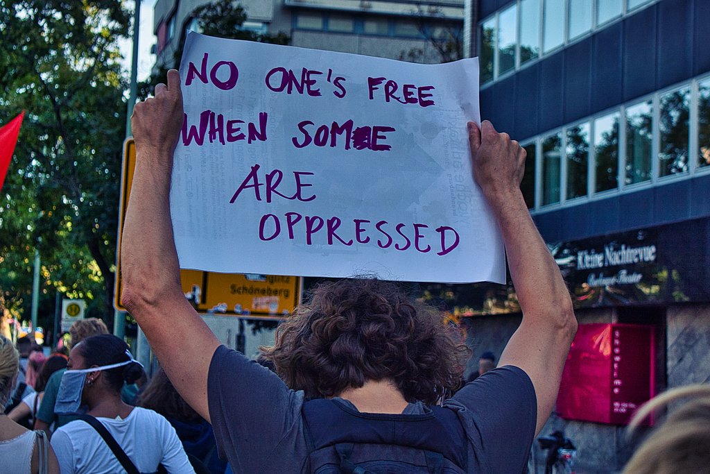 Demo von hinten fotografiert. Eine Person hält ein Schild hoch, auf dem steht: No one's free if some are oppressed.